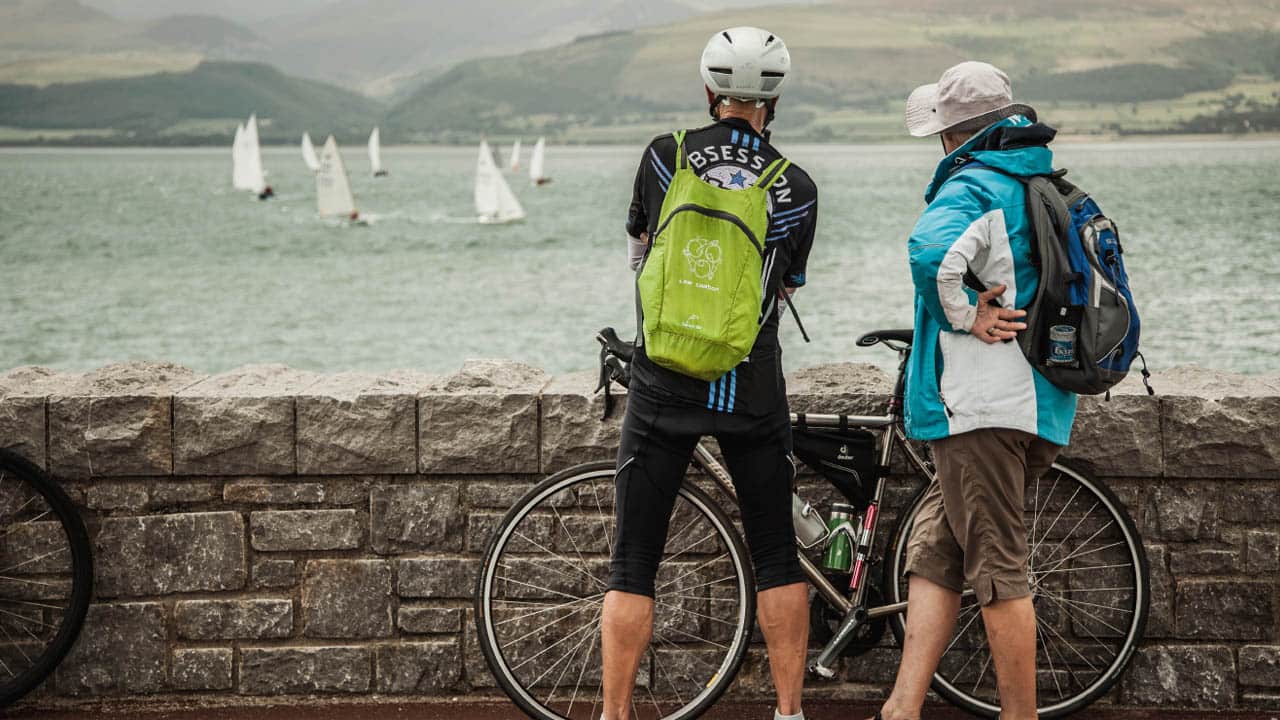 Two men with bikes standing by a wall looking out to sea at the sailboats